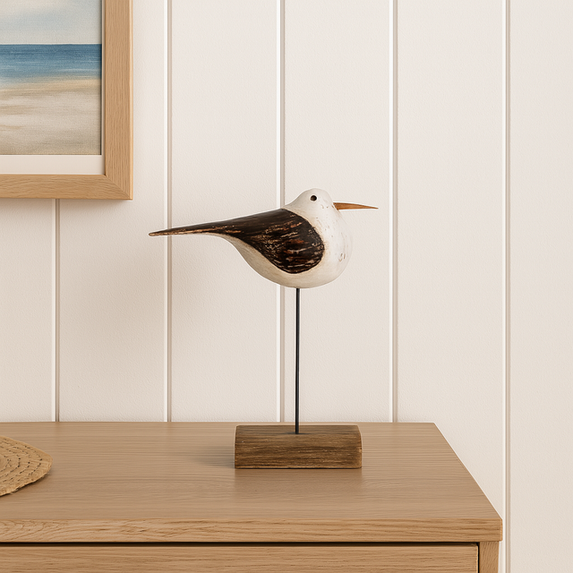 Dekorativ trebåt-måke på stativ står på lyst trebord mot hvit panelvegg, med maritimt bilde i ramme ved siden av.