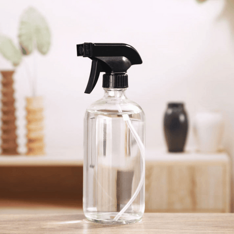 Klar glassprayflaske med svart spraytopp og hvit slange, stående på bord i stilren og naturlig innredet stue.