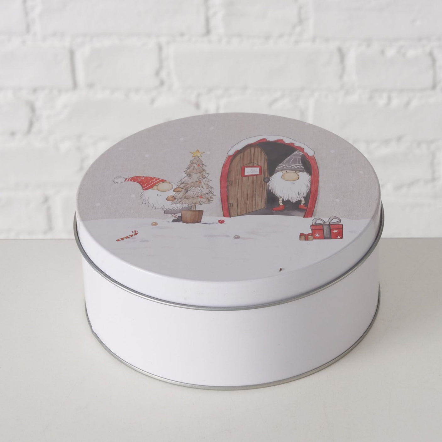 Hvit kakeboks med julemotiv av nisse med juletre og nisse ved dør, mot lys mursteinsvegg.