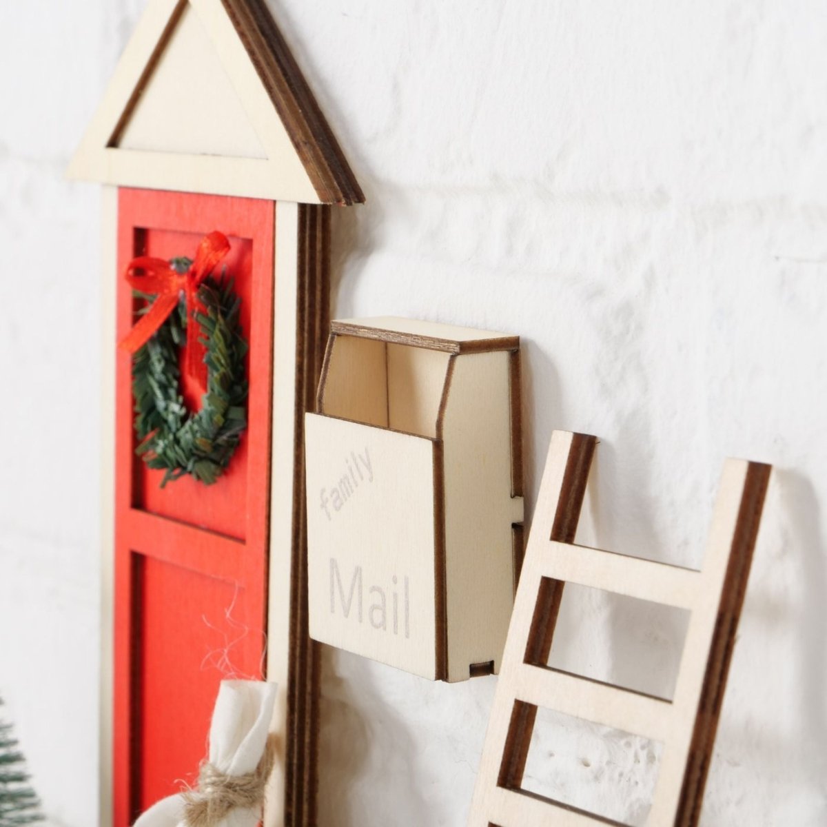 Rød miniatyrdør med julekrans, hvid postkasse og stige på væggen.