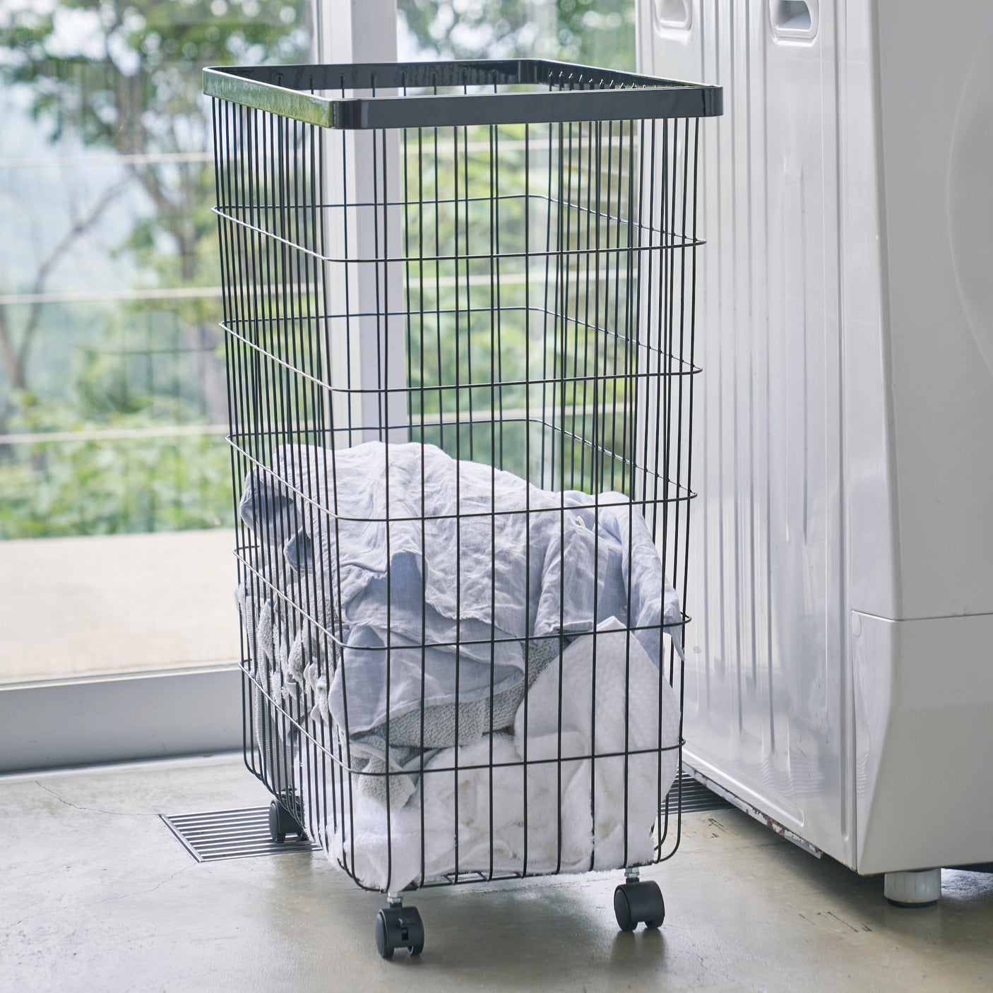 Praktisk skittentøyskurv på hjul i sort metall, plassert i et lyst rom med grønne planter.