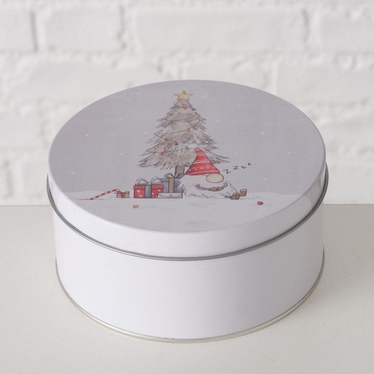 Hvid kageboks med julemotiv med en sovende nisse ved et juletre og gaver, mod en lys mursteinvæg.