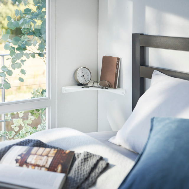Hvit veggmontert hjørnehylle brukt som nattbord ved siden av en seng, med klokke, bok og briller på toppen.