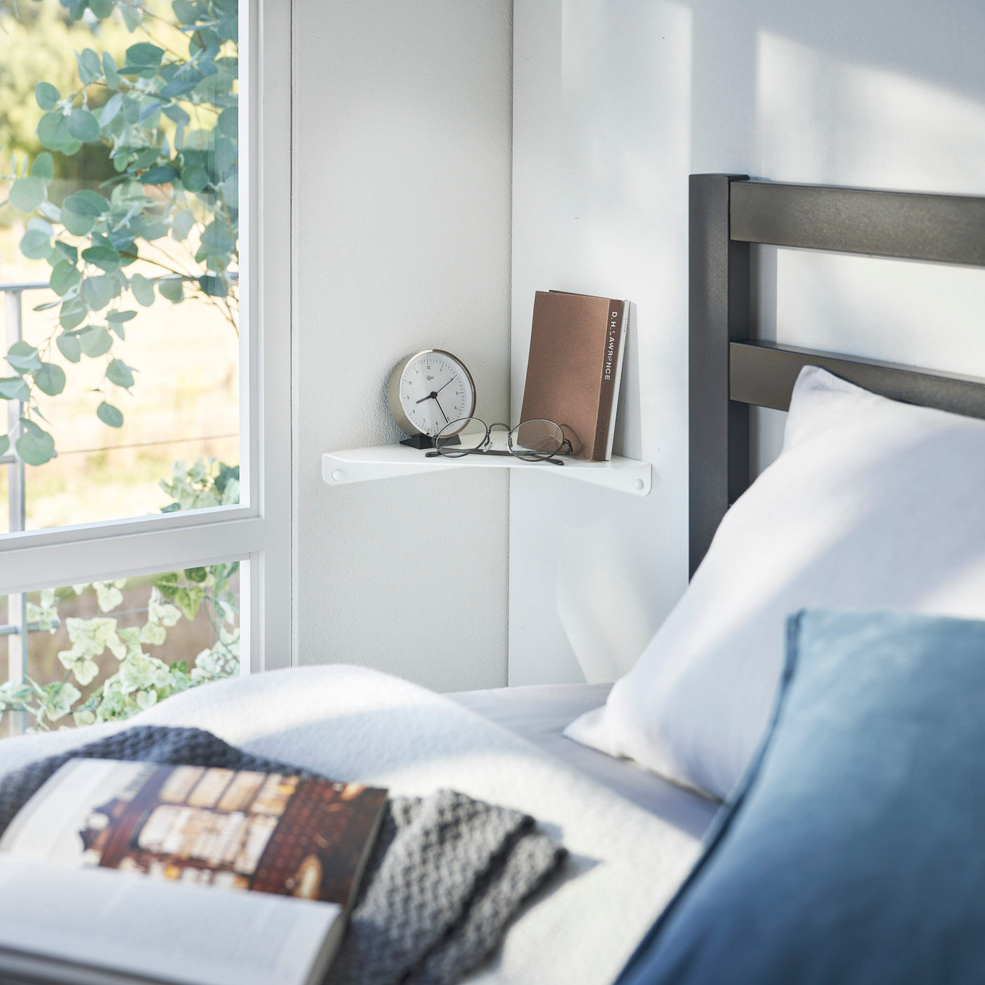 Hvit veggmontert hjørnehylle brukt som nattbord ved siden av en seng, med klokke, bok og briller på toppen.