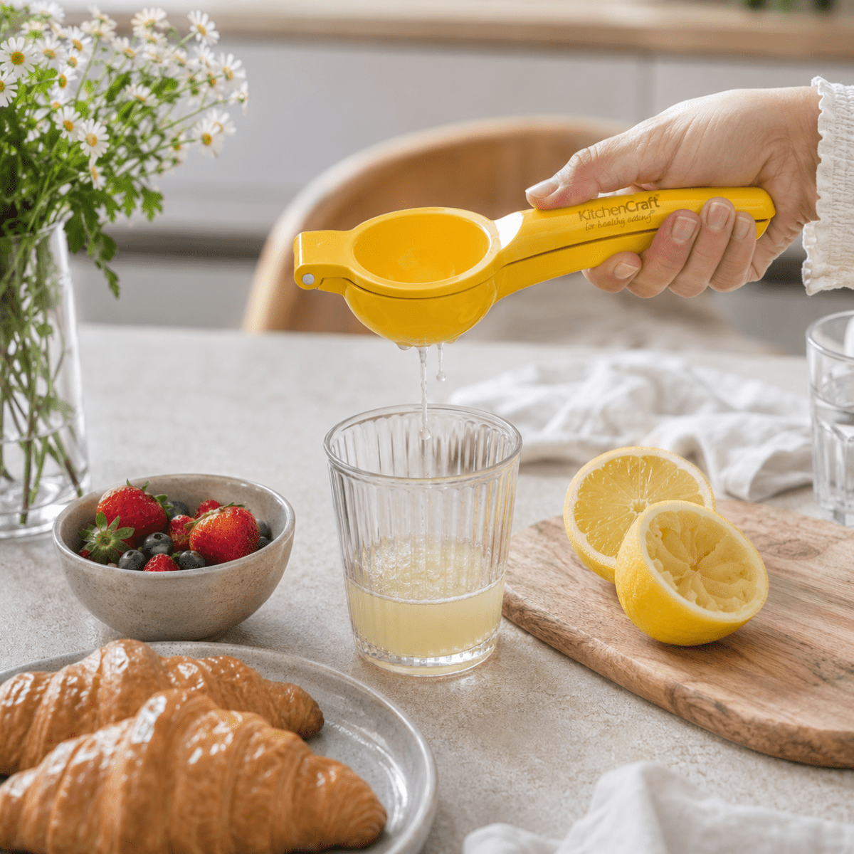 Hånd som holder en gul KitchenCraft-sitronpresse over et glass med sitronsaft på et lyst frokostbord. - Lindas dekor