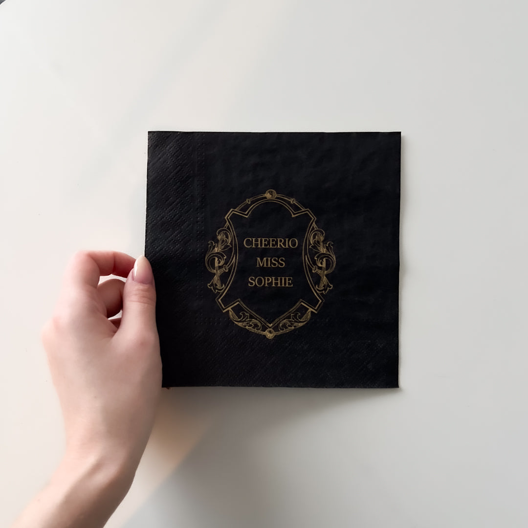 Hånd holder sort serviett med gulltrykk "Cheerio Miss Sophie" på hvit bakgrunn.