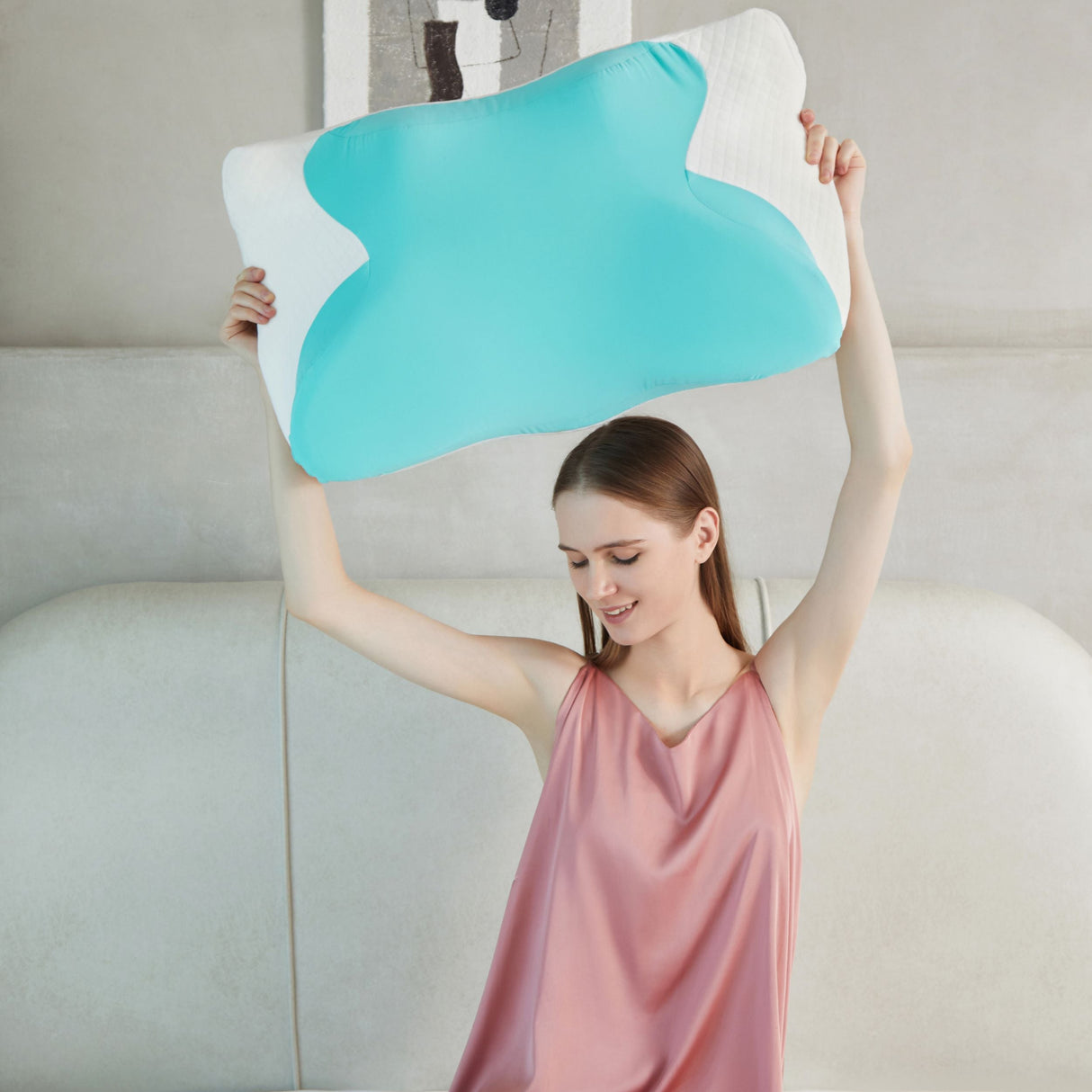 Kvinne i rosa nattkjole smiler og løfter en blå og hvit ergonomisk minneskumpute over hodet.