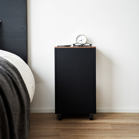 Sidebord i sort metall med bordplate i tre, plassert ved seng med lampe på toppen.