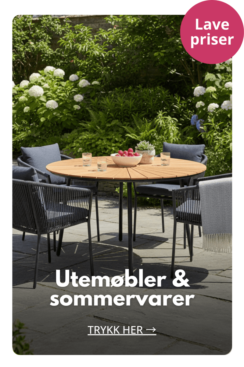 Hagegruppe med rundt spisebord og stoler på terrasse omgitt av grønne planter og sommerblomster