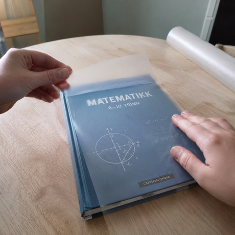 Transparent matt kontaktplast lagt over norsk skolebok i matematikk for å beskytte omslaget.