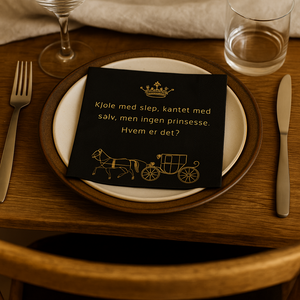 Svart serviett med gullmotiv og tekst ligger realistisk på tallerken i naturlig lys.
