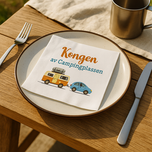 Serviett med teksten «Kongen av Campingplassen» ligger på tallerken på sommerlig trebord utendørs.
