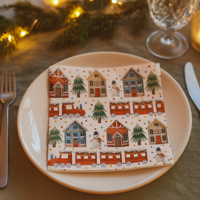 Serviett med julemotiv av tog, snømenn og hus ligger på tallerken i julemiljø.
