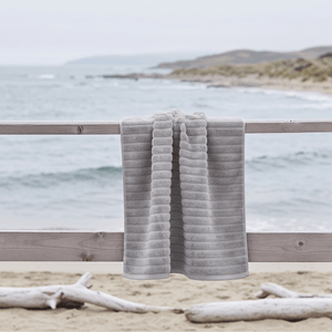 Grått Inu badehåndkle fra Zone Denmark lagt over strandstol ved vannet i rolig kystmiljø.