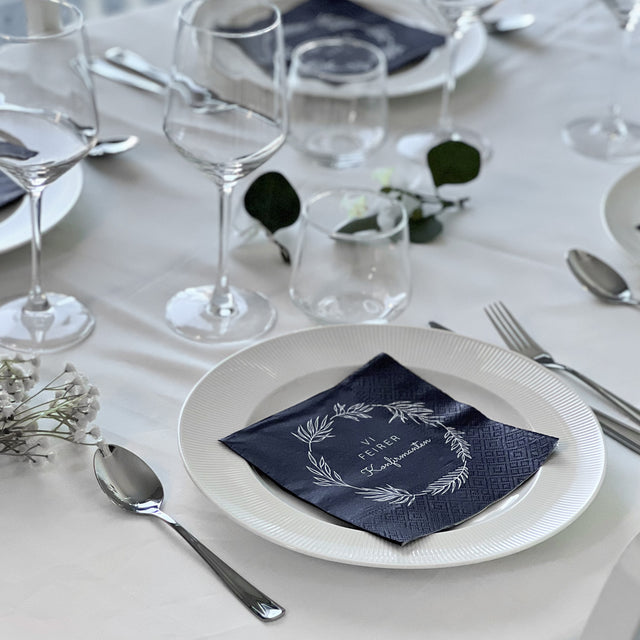 Mørkeblå serviett med teksten «Vi feirer konfirmanten» ligger på tallerken i elegant borddekking.
