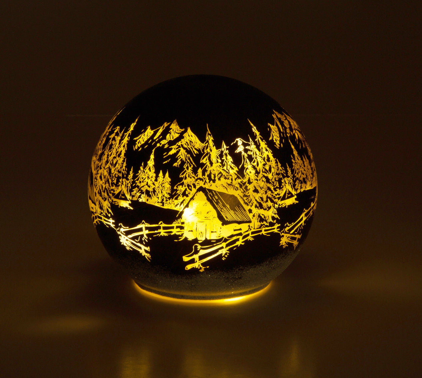 Rund dekorlampe med inngravert motiv av hytte, fjell og skog i varmt lys.