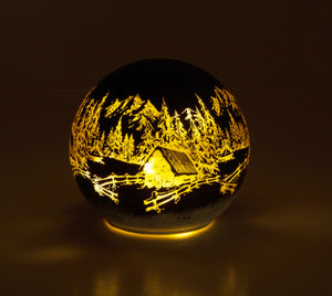 Rund dekorlampe med inngravert motiv av hytte, fjell og skog i varmt lys.