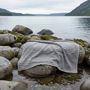 Grått Inu badehåndkle fra Zone Denmark lagt over steiner ved vannet i nordisk naturmiljø.