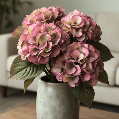 Nærbilde av kunstig hortensia i rosa toner med realistiske blader, plassert i grå keramikkvase på bord i stilren og lun stue.